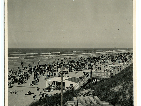 Ein Foto von einem Strand mit vielen Strandkörben und Menschen, die liegen, baden oder rumlaufen. Im Hintergrund ist das Meer zu sehen und im Vordergrund eine Sanddüne mit einer Treppe und einem Schild mit der Aufschrift »Verboden voor Joden«.