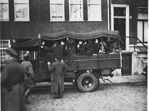 Ein überdachter Wagen auf einer Straße, darauf Personen, im Vordergrund Uniformierte.