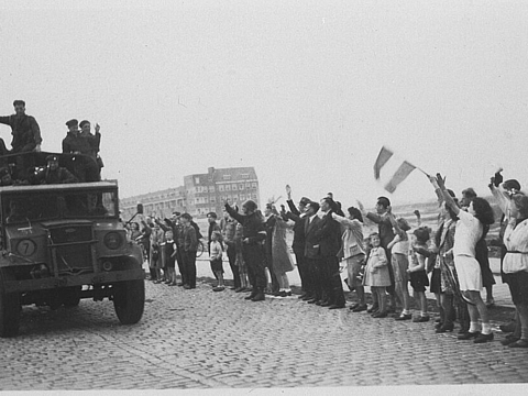 Menschen mit Fahnen stehen winkend am Straßenrand, ein Militärfahrzeug fährt an ihnen vorbei.