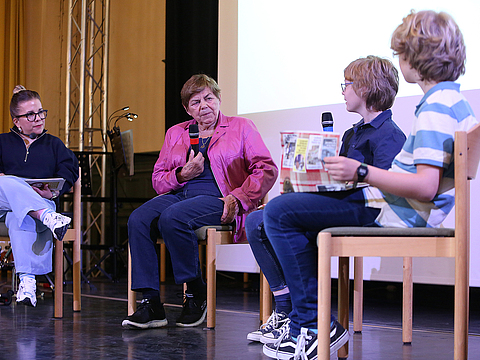 Vier Menschen sitzen nebeneinander auf einer Bühne. Sie halten Mikrofone in den Händen und sehen einander an.