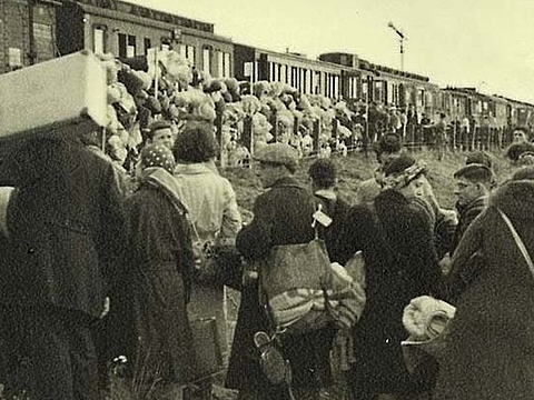 Viele Menschen in Mänteln mit Gepäck stehen gedrängt in einer Schlange vor einem Güterzug.