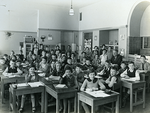 Ein Foto von einem Klassenraum mit Pulten. Im Vordergrund sitzen Schüler*innen, im Hintergrund stehen sie und schauen in die Kamera.