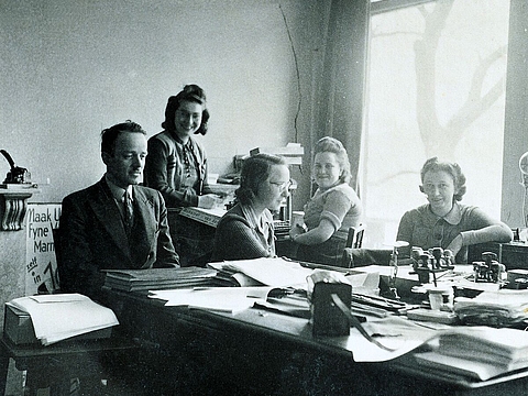 Fünf Personen sitzen an Schreibtischen, darauf liegen Bücher und Papiere.