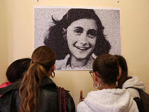 An einer Wand hängt ein schwarz-weiß Porträt eines jungen, lächelndes Mädchens. Vier Schülerinnen stehen mit dem Rücken zur Kamera und betrachten das Porträt.