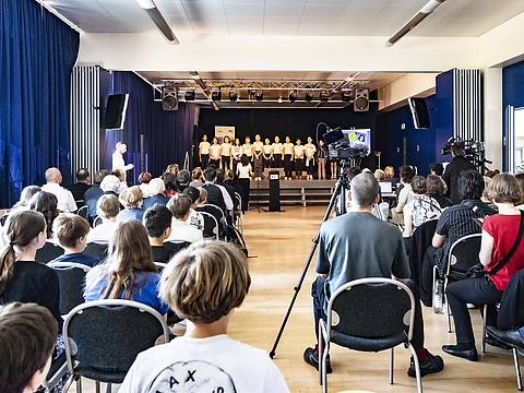 Auf dem Foto ist ein langer Saal zu sehen mit einer Bühne, auf dem viele Kinder stehen. Rechts und links vorne im Saal stehen viele Stühle, auf denen Menschen sitzen. Die sitzenden Menschen sind mit dem Rücken zur Kamera.