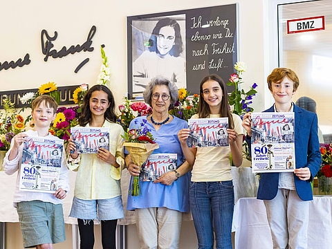 Auf dem Foto sind fünf Menschen zu sehen, die nebeneinander aufgereiht sind und alle eine Zeitung in der Hand haben, die sie in die Kamera zeigen. Die Person in der Mitte ist eine ältere Frau und die jeweils neben ihr stehenden Personen sind Kinder.