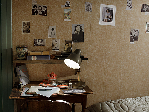Ein Foto von einem Ausschnitt eines Zimmers. Im Zimmer stehen ein schmaler Schreibtisch und davor ein Stuhl aus Holz. Auf dem Schreibtisch liegen Bücher, ein Stift, eine Schale mit Erdbeeren und eine eingeschaltete Tischlampe. An der Wand hinter dem Tisch kleben Fotos und Ansichtskarten. An der rechten Seite des Fotos ist die Kante eines Betts zu sehen.