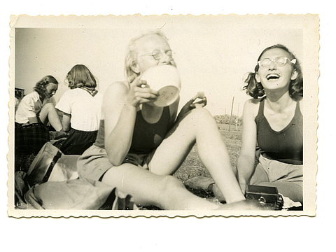 Ein Foto von einem Bild von zwei jungen Frauen, die auf einer Wiese sitzen. Die eine junge Frau hält eine weiße Schüssel in der Hand. Im Hintergrund sitzen zwei weitere junge Frauen.