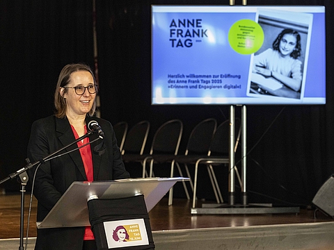 Auf dem Foto ist eine Frau zu sehen, die an einem Rednerpult in ein Mikrofon spricht. Sie steht vor einer Bühne. Hinten rechts ist ein Bildschirm, auf dem ein Bild eingeblendet ist.