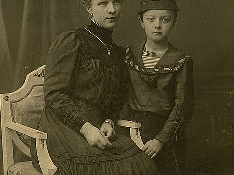 Eine Frau auf einem Stuhl, daneben ein Kind, beide gut gekleidet.